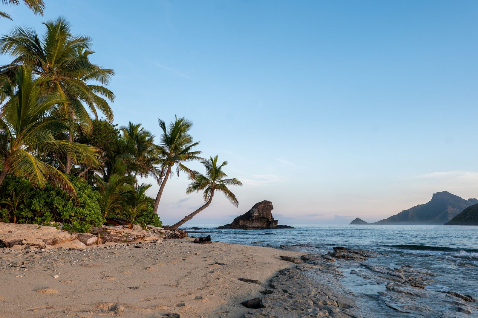 Palmbomen en een rotseilandje aan een strand in Fiji bij zonsopkomst